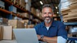 © Lens Legacy - A man with gray hair and beard smiling while working on a laptop computer inside a warehouse filled with shelves and boxes, projecting a professional attitude.