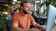 © Lens Legacy - A bearded man in an orange shirt is seated at an office desk, typing on his computer with focused attention, surrounded by colleagues and common modern office decor.