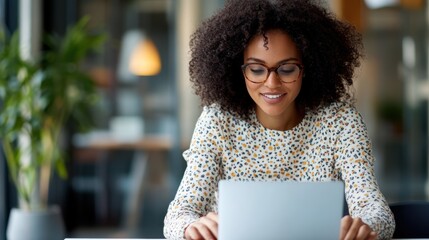 A focused woman with curly hair and glasses sits at her laptop in a modern office, exuding dedication and concentration in a bright and lively setting.