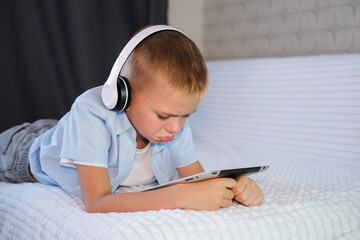 Boy plays games on tablet, lies on sofa. Laptop, home education. Knowledge website, educational software and small child broadcasting youth development lesson on headphones.
