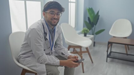 Sticker - A young man in medical white coat using smartphone in a modern clinic waiting room.