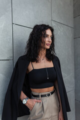 Wall Mural - Glamour fashion curly beautiful woman in black clothes with blazer and top with pants stands near a gray wall on the street