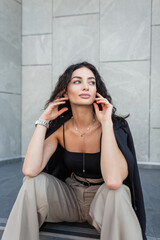 Wall Mural - Fresh beautiful glamorous fashion woman with curly hairstyle in fashionable elegant clothes with blazer, top and pants sitting near gray building in city