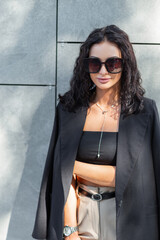 Wall Mural - Pretty curly fashion urban woman with sunglasses  in elegant clothes with blazer, top and pants stands near a gray wall