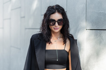 Wall Mural - Beautiful young brunette elegant curly woman with vintage sunglasses in black blazer with top on gray background in city