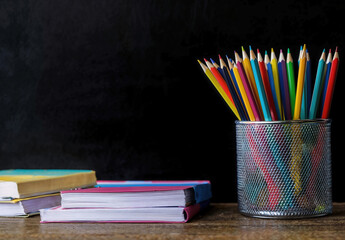 Poster - Colorful pencils in a metal container with books on a wooden table against a blackboard background