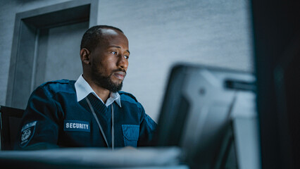 African American male security operator or dispatch in uniform uses tablet computer, looks at screens sitting at the workplace in monitoring center during night shift. Surveillance system and CCTV.