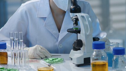 a scientist is looking into a microscope for research. on the table are lab glassware containing tes