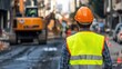 © 69 - An engineer in a safety vest and helmet, working on a construction site with heavy machinery in the background, overseeing roadwork operations.