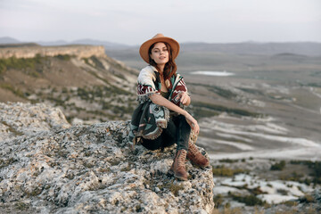 Wall Mural - Woman in cozy attire sitting on mountain rock with hat and scarf, enjoying peaceful solitude