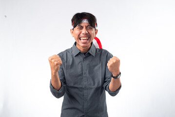 Indonesian man celebrating with fists raised, wearing a grey shirt and a red-and-white headband. He is smiling broadly, showing excitement and national pride, captured in a festive and joyful moment.