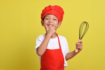 Cute little chef holding kitchen whisk. Asian boy wearing red apron and hat on isolated yellow background