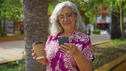 Poster - Beautiful mature woman with grey hair and glasses enjoying coffee while using her smartphone in an urban park on a sunny day