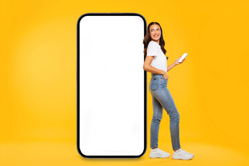 Happy young European lady standing by huge smartphone with white blank screen, showing online offer, yellow studio background, mockup