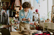 © AnnaStills - Young man standing in cluttered art studio bending over smartphone concentrating on screen focusing on his work surrounded by various paints. Shelves are filled with art supplies and decorations