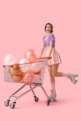 Poster - Happy young fashionable woman in headphones with shopping cart and balloons on pink background