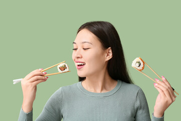 Poster - Beautiful young Asian woman holding tasty sushi rolls with chopsticks on green background