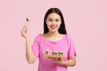 Wall Mural - Beautiful young Asian woman holding chopsticks and wooden tray with tasty sushi rolls on pink background