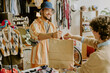 © AnnaStills - Smiling man wearing glasses and a hat handing shopping bag in a thrift shop with various clothes hanging in background