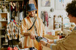 © AnnaStills - Smiling man wearing hat and glasses making payment at clothing store counter with cashier accepting card. Shelves filled with garments and decorations visible in background