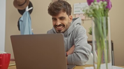 Wall Mural - Happy hispanic man with beard using laptop in a cozy home interior, expressing excitement and success.