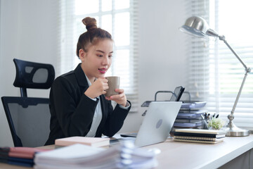 A confident businesswoman in a black suit is sitting at her desk, smiling as she works on her laptop, showcasing a modern and professional workspace