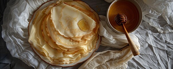 A stack of golden brown crepes on a white plate with a bowl of honey and a honey dipper.