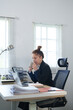 © snowing12 - A confident businesswoman in a black suit is sitting at her desk, smiling as she works on her laptop, showcasing a modern and professional workspace