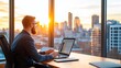 © Pual GotoMars - A man sits at a desk, working on his laptop, while the sun sets over the city skyline through a large window.