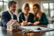 © jex - Close-up of a miniature house model and keys, with a real estate agent and a couple signing a contract in the blurred background.