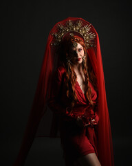 Wall Mural - dramatic close up portrait a female model wearing a red silk dress and veil robes, with a ornate golden fantasy crown headdress. isolated on a dark studio background with  silhouette rim lighting 