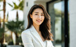 © Surachetsh - An Asian woman in a white suit smiles confidently while posing outdoors, with a backdrop of modern architecture and office buildings.
