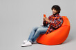 © ViDi Studio - Full body young happy smiling Indian man wears red shirt casual clothes sit in bag chair hold in hand use mobile cell phone isolated on plain grey color background studio portrait. Lifestyle concept.