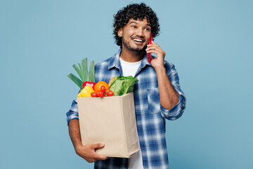 Young Indian man he wear shirt hold brown craft bag for takeaway mock up with food products talk speak on mobile cell phone isolated on plain blue background. Delivery service from shop or restaurant.