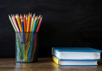 Aufkleber - Colorful pencils in a cup are standing near books on a wooden desk with a blackboard in the background, providing copy space