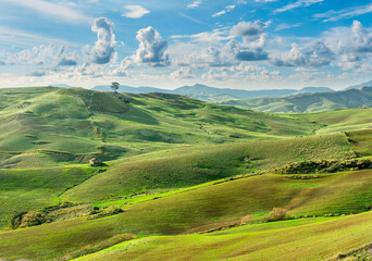 beautiful rural view at summer or spring season fields and hills with rustic grassland and nice mountains with blue cloudy sky on background of countyside landscape