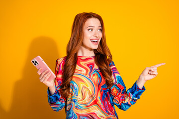 Wall Mural - Photo portrait of lovely young lady hold device point look empty space dressed stylish colorful garment isolated on orange color background