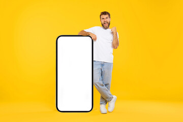Excited guy leaning on big smartphone with empty white screen and shaking clenched fist, celebrating win, standing over yellow background, mockup