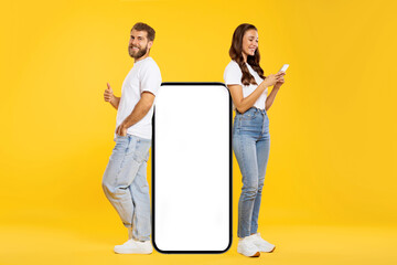 Wall Mural - Man and woman standing near huge cellphone with blank screen, lady texting on phone while man showing thumb up and smiling, mockup