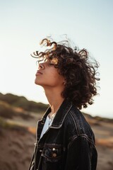 A person with curly hair and wearing a denim jacket looking away, standing outdoors on a sunny day with a natural landscape in the background.