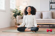 © kenchiro168 - Relaxed African American woman meditating in seated position on yoga mat at home, listening to music with headphones.
