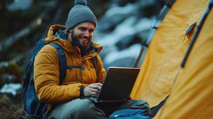 A joyful camper engages with his laptop beside a cozy yellow tent in a tranquil mountainous landscape as dusk approaches