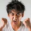 © Nina - Young man screams to expressing intense frustration with clenched fists and a fierce expression against a plain background. Japanese man shows anger, with his fists raised and a fierce grimace on face