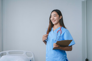 Young female doctor smiles warmly in a hospital room, holding a clipboard and wearing blue and red scrubs, radiating professionalism and care