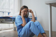 © amnaj - Exhausted young asian female nurse in blue uniform sitting on the floor near the bed and touching her head while suffering from headache