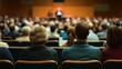 © venusvi - Audience seen from the back, engaged in a lecture in a well-lit conference hall with a speaker addressing the crowd