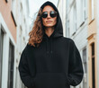 © Pemika - A stylish woman in a black hoodie and sunglasses confidently poses on a city street, showcasing urban fashion and attitude.