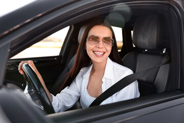 Wall Mural - Smiling young woman in sunglasses with seatbelt driving car, view from outside