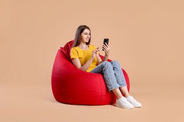 Smiling woman using smartphone while sitting on red bean bag chair against beige background