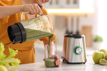 Poster - Woman pouring fresh smoothie from blender cup into glass at white marble table in kitchen, closeup